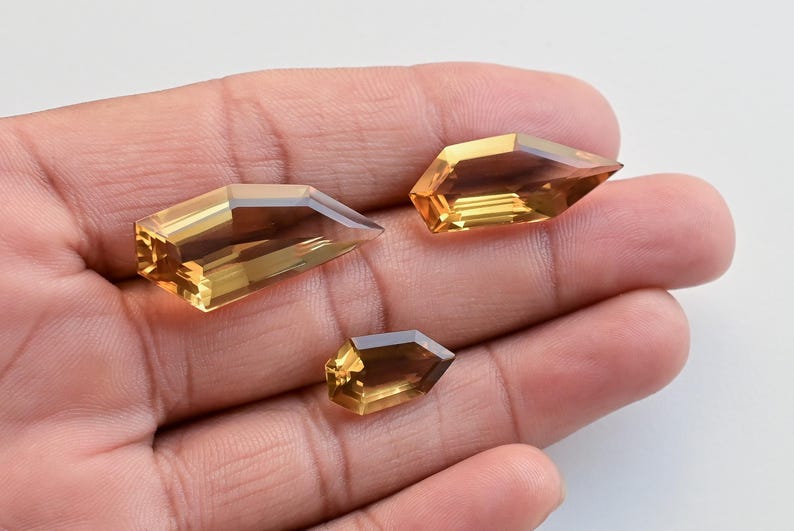 May include: Three faceted, golden-yellow gemstones with pointed ends are displayed in a hand. The gems have a rectangular shape with angled sides, catching the light to reveal their internal structure. The gems vary in size.