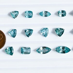 May include: An assortment of aquamarine gemstones in various shapes, including round, square, and marquise cuts. A U.S. penny is included for size comparison. The gemstones are a light blue-green color and are displayed on a white surface.