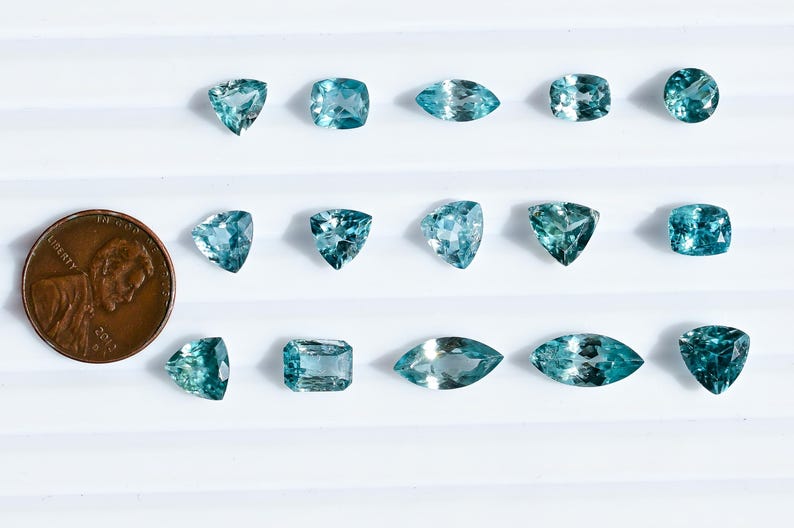 May include: An assortment of aquamarine gemstones in various shapes, including round, square, and marquise cuts. A U.S. penny is included for size comparison. The gemstones are a light blue-green color and are displayed on a white surface.