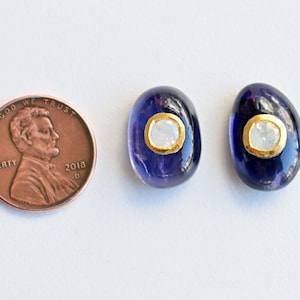 May include: A pair of oval-shaped earrings with a dark purple stone and a small white stone set in a gold bezel. The earrings are about the size of a UK penny.