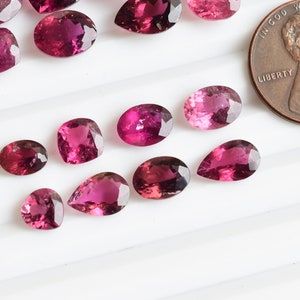 May include: A collection of pink and purple gemstones, some are pear shaped and others are oval. The gemstones are arranged on a white surface.  A US penny is included for scale.