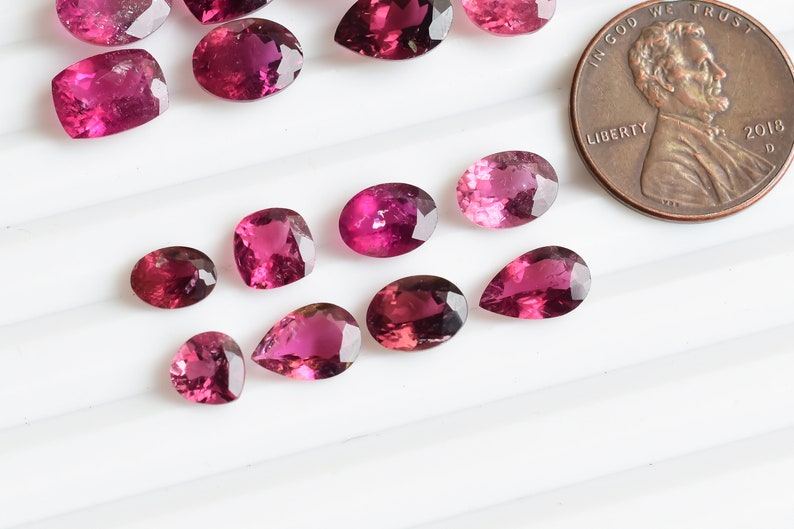 May include: A collection of pink and purple gemstones, some are pear shaped and others are oval. The gemstones are arranged on a white surface.  A US penny is included for scale.