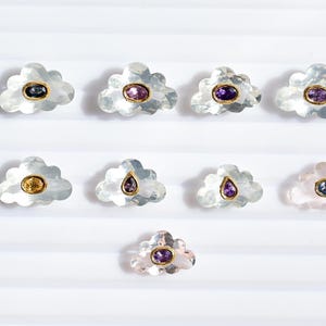 Peut inclure: Une collection de bijoux en forme de nuage avec diverses pierres précieuses de couleur serties dans de l'or. Les pierres précieuses comprennent des teintes violettes, bleues et jaunes. Les bijoux sont exposés sur une surface blanche texturée.