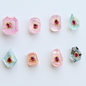 May include: Eight gemstone pendants in various shapes and colours, including pink, peach, and teal. Each pendant features a central red gemstone set in a gold-coloured bezel. The gemstones are arranged on a white background.