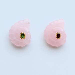 May include: A pair of light pink, seashell-shaped earrings. Each earring features a gold setting with a central oval-shaped gemstone. One earring has a green gemstone, the other a red gemstone.