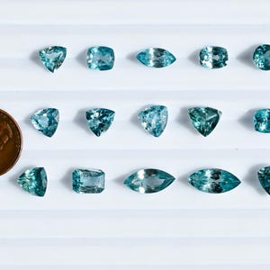 May include: An assortment of teal-colored gemstones in various shapes, including triangles, squares, and ovals, are displayed on a white surface. A U.S. penny is included for size comparison. The gemstones are likely for jewelry making.