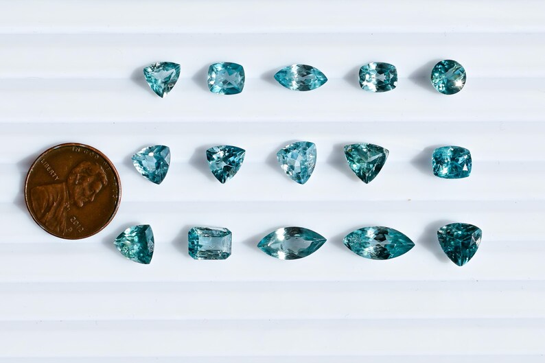 May include: An assortment of teal-colored gemstones in various shapes, including triangles, squares, and ovals, are displayed on a white surface. A U.S. penny is included for size comparison. The gemstones are likely for jewelry making.