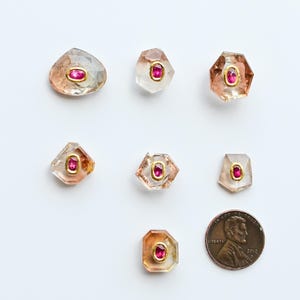 May include: Nine gemstone crystals with a penny for scale. The crystals have a clear, peach, and gold color with a small, oval, red gemstone in the center. The crystals are various shapes, including oval, hexagonal, and irregular.
