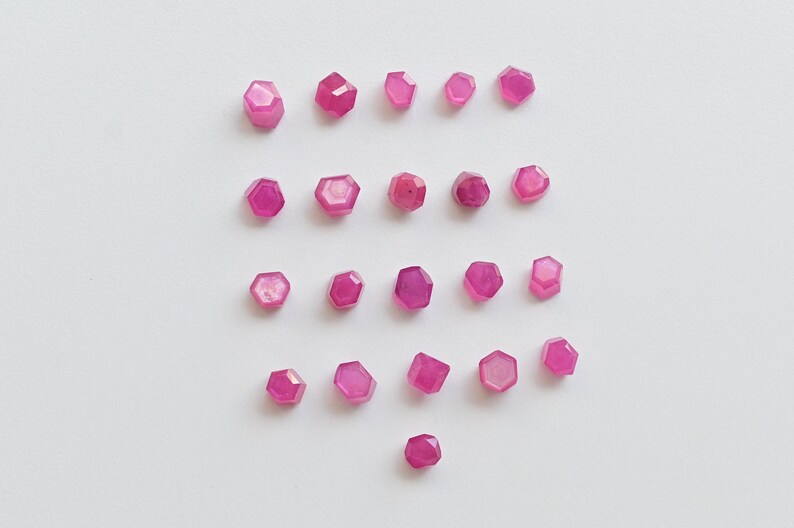 May include: Twenty-one pink faceted gemstones arranged in four rows on a white background. The gemstones are hexagonal in shape.