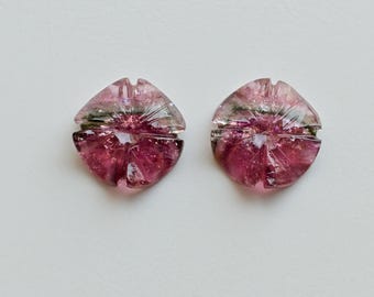 May include: Two faceted, pink and clear tourmaline gemstones. The gemstones have a flower-like shape with a central starburst pattern and are set against a white background. The stones have a mix of pink and green inclusions.