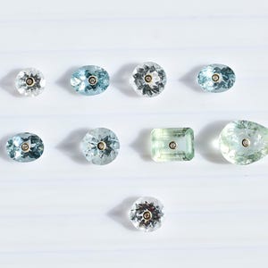 May include: A collection of gemstones in different shapes and hues, including oval, round, and pear-cut. The gems are in shades of blue and green, with a small gold accent in the centre of each stone. The gems are arranged on a white surface.