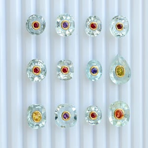 May include: A collection of gemstone cabochons in diverse shapes, including round, oval, and teardrop. Each gem features a central, colourful stone, such as red, purple, or yellow, set in a gold bezel. The gems are displayed on a white, ridged surface.