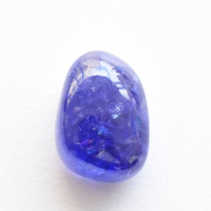 K&ouml;nnte beinhalten: Ein polierter, oval geformter Tansanit-Edelstein. Der Edelstein hat ein tiefes, sattes Blau mit violetten Nuancen und schillernden Reflexen. Er liegt vor einem schlichten wei&szlig;en Hintergrund, der seine Farbe und Klarheit hervorhebt.