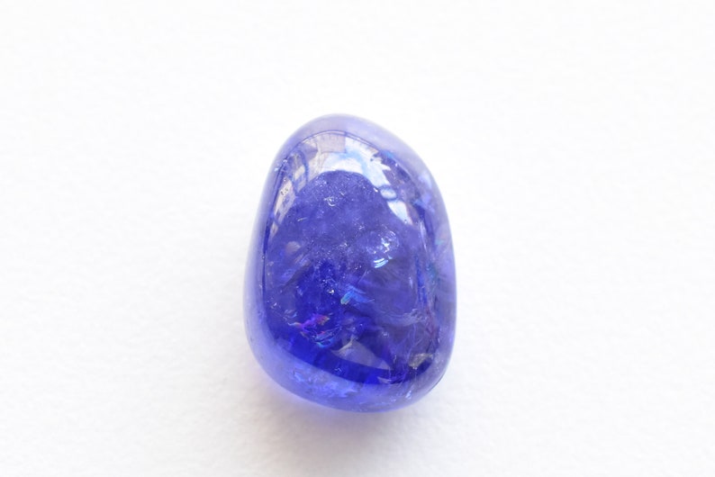 K&ouml;nnte beinhalten: Ein polierter, oval geformter Tansanit-Edelstein. Der Edelstein hat ein tiefes, sattes Blau mit violetten Nuancen und schillernden Reflexen. Er liegt vor einem schlichten wei&szlig;en Hintergrund, der seine Farbe und Klarheit hervorhebt.
