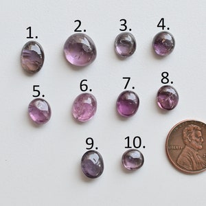 May include: Ten purple and pink oval-shaped gemstones, numbered 1 through 10, are arranged in two rows on a white surface. A UK penny is included for size comparison.