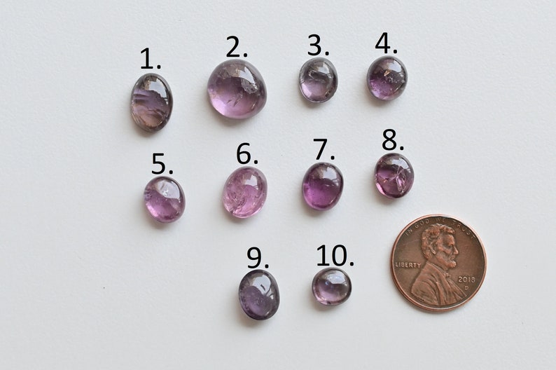 May include: Ten purple and pink oval-shaped gemstones, numbered 1 through 10, are arranged in two rows on a white surface. A UK penny is included for size comparison.