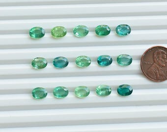 May include: A collection of 15 oval-shaped green gemstones arranged in three rows of five. The gemstones are various shades of green, ranging from light to dark. A US penny is included for scale.