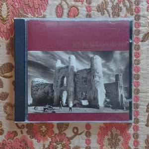May include: A CD case featuring the album "The Unforgettable Fire" by U2. The cover art displays a black and white image of a castle ruin under a cloudy sky. The top of the case has a burgundy color with the album title.