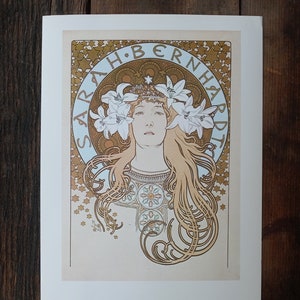 May include: A vintage poster featuring a woman with long flowing hair, wearing a crown of lilies. The poster is in Art Nouveau style and features a gold and brown color scheme. The text "SARAH BERNHARDT" is written in a decorative font around the woman's head.