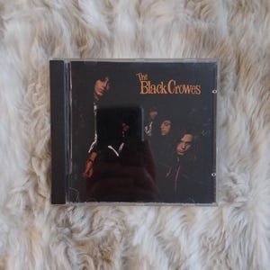May include: A CD case featuring the band The Black Crowes. The album cover is predominantly black, with the band's name in orange lettering. The image shows the band members in a dark, moody setting.