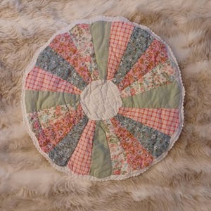 May include: A round, handmade patchwork cushion with a scalloped edge. The cushion features a sunburst design with alternating segments of floral, gingham, and solid fabrics in pink, blue, and green. The center is white.