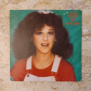 May include: Vintage album cover featuring Gilda Radner with the text "Gilda Radner Live From New York." The cover has a green background with a portrait of Radner wearing a red shirt and white overalls. The album cover is square.