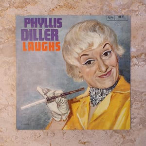 May include: Vintage vinyl record album cover featuring Phyllis Diller. The album cover has the text "PHYLLIS DILLER LAUGHS" in purple and orange. The cover art is a painted portrait of Phyllis Diller in a yellow jacket, holding a cigarette holder.