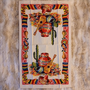 May include: White rectangular kitchen towel with a red border and a colorful Mexican-themed design. The design includes a sombrero, guitar, cactus, and a decorative vase. The towel has a repeating pattern of colorful stripes.