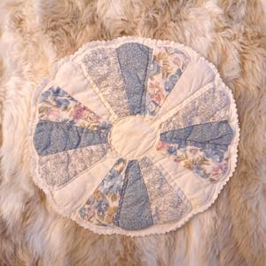 May include: A round, quilted cushion with a scalloped edge. The cushion features alternating sections of floral and patterned fabric in shades of blue, pink, and white. The center is a solid off-white fabric.