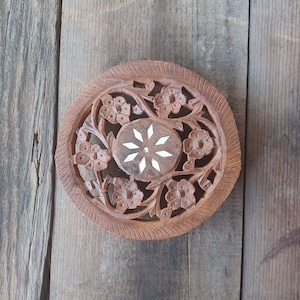 May include: A round wooden coaster with intricate floral carvings and a white starburst design in the center.