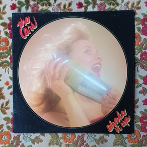 May include: The Cars "Shake It Up" album cover. A woman with blonde hair holds a cocktail shaker to her ear, mouth open in a shout. The album title is in red and green, with the band's name in green.
