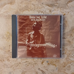 May include: A CD case featuring the album cover for "Hound Dog Taylor and the HouseRockers." The cover art is sepia-toned, showing a man playing a guitar. The album title is printed in a bold, serif font.