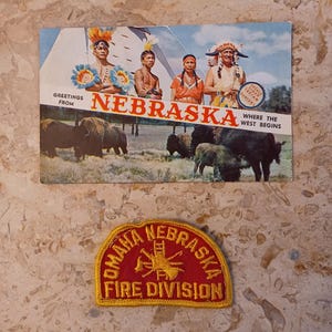 May include: Vintage Nebraska postcard featuring Native American figures and bison, with the text "Greetings from Nebraska" and "Where the West Begins." Below is a red and yellow Omaha Nebraska Fire Division patch.