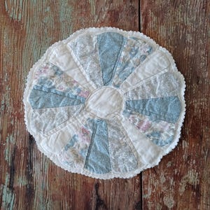 May include: A circular quilted fabric mat featuring a central white circle surrounded by alternating wedge-shaped panels. The panels display various floral and botanical patterns in shades of dusty blue, sage green, and soft pink, finished with a scalloped white edge.
