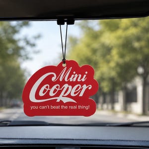 May include: A red car air freshener hanging from a rearview mirror. The air freshener is shaped like a classic soda logo and features the white text Mini Cooper and the phrase you can't beat the real thing! printed on the front surface.