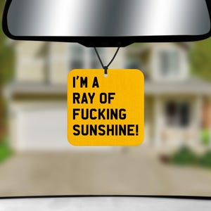 May include: A square yellow car air freshener hanging from a rearview mirror by a black string. The air freshener features the black text I'M A RAY OF FUCKING SUNSHINE! printed in a bold, sans-serif font centred on the bright yellow background.