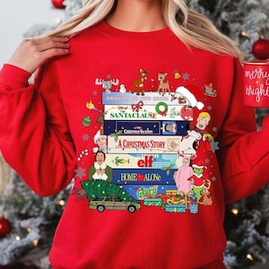 May include: Red Christmas sweater with a stack of classic Christmas movie VHS tapes, including 'Home Alone', 'Elf', 'A Christmas Story', 'National Lampoon's Christmas Vacation', 'The Santa Clause', and 'Rudolph the Red-Nosed Reindeer'.