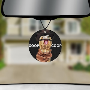 May include: A circular hanging car air freshener featuring a graphic of a peanut character wearing a brown cowboy hat and scarf. The character has a wide-open mouth and eyes, with the text GOOP printed on the left and right sides in white.