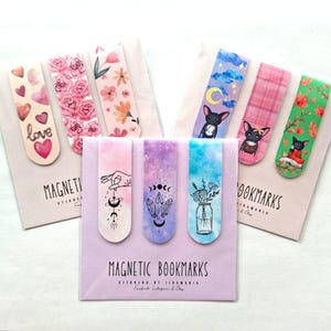 Puede incluir: Una colección de marcadores magnéticos con varios diseños. Algunos presentan patrones florales, corazones y la palabra "love". Otros muestran un perro, motivos celestiales e ilustraciones de acuarela. Los marcadores están empaquetados en cartulina rosa claro y morada.