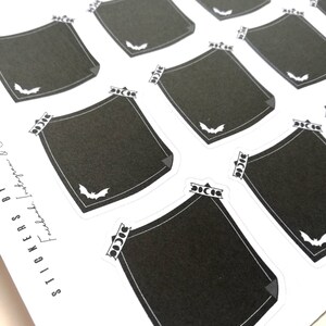 May include: Ten black and white Halloween themed stickers with a bat design. Each sticker has a black square with a white border and a white bat in the bottom right corner. The stickers are on a white background.
