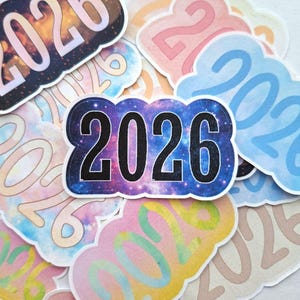 Puede incluir: Una colección de pegatinas coloridas con el año "2026" impreso. Las pegatinas presentan una variedad de diseños, incluyendo un patrón de galaxia, colores pastel y una forma de nube. Los números están en negrita, en negro.