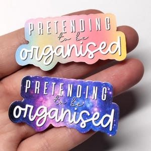 Puede incluir: Dos pegatinas con la frase "PRETENDING to be organised" en escritura blanca. Una pegatina tiene un fondo degradado colorido, y la otra tiene un fondo con temática de galaxia con tonos morados, azules y rosas.