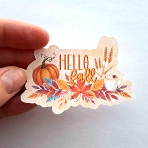 May include: A fall-themed sticker with watercolour illustrations. It depicts a pumpkin, autumn leaves, wheat, and a coffee cup. The words "Hello Fall" are written in orange script.
