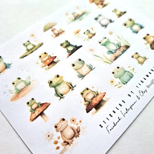 May include: Sheet of stickers featuring watercolor illustrations of frogs in various poses. The frogs are in shades of green, brown, and beige, some sitting on lily pads or mushrooms. The stickers have a whimsical, nature-inspired theme.