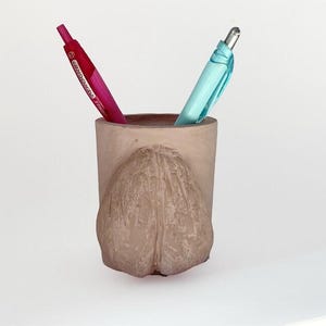 May include: A beige ceramic pen holder with a textured, rounded front design. Two pens are inside: one is pink and the other is light blue. The pen holder is on a white surface.