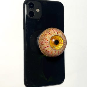 May include: A black smartphone with a pop-up phone grip featuring a realistic eyeball design. The eyeball has a yellow iris, a black pupil, and red veins. The phone is on a white background.