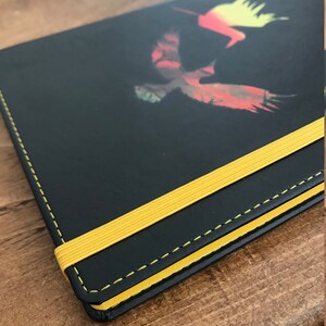 A black A5 faux leather notebook with a yellow elastic closure, yellow edged paper, and yellow stitching.  The notebook has a design in the middle of two hummingbirds, one yellow and orange ombre and the other green and brown and red.