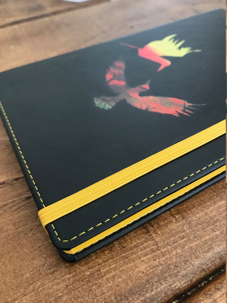 A black A5 faux leather notebook with a yellow elastic closure, yellow edged paper, and yellow stitching.  The notebook has a design in the middle of two hummingbirds, one yellow and orange ombre and the other green and brown and red.