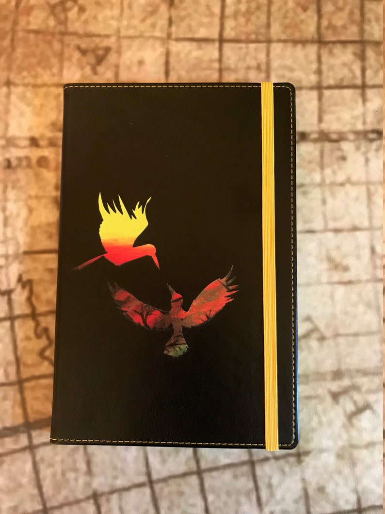 A black A5 faux soft touch leather notebook with a yellow elastic closure and yellow stitching.  The notebook has a design in the middle of two hummingbirds, one yellow and orange ombre and the other green and brown and red.