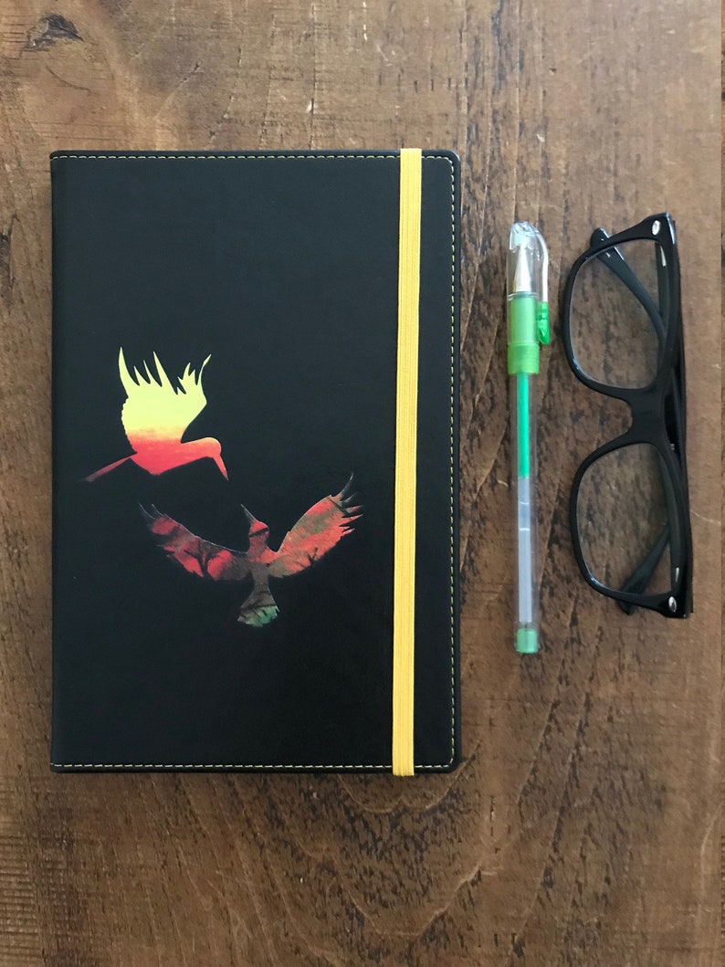 A black A5 faux leather notebook with a yellow elastic closure, yellow edged paper, and yellow stitching.  The notebook has a design in the middle of two hummingbirds, one yellow and orange ombre and the other green and brown and red.